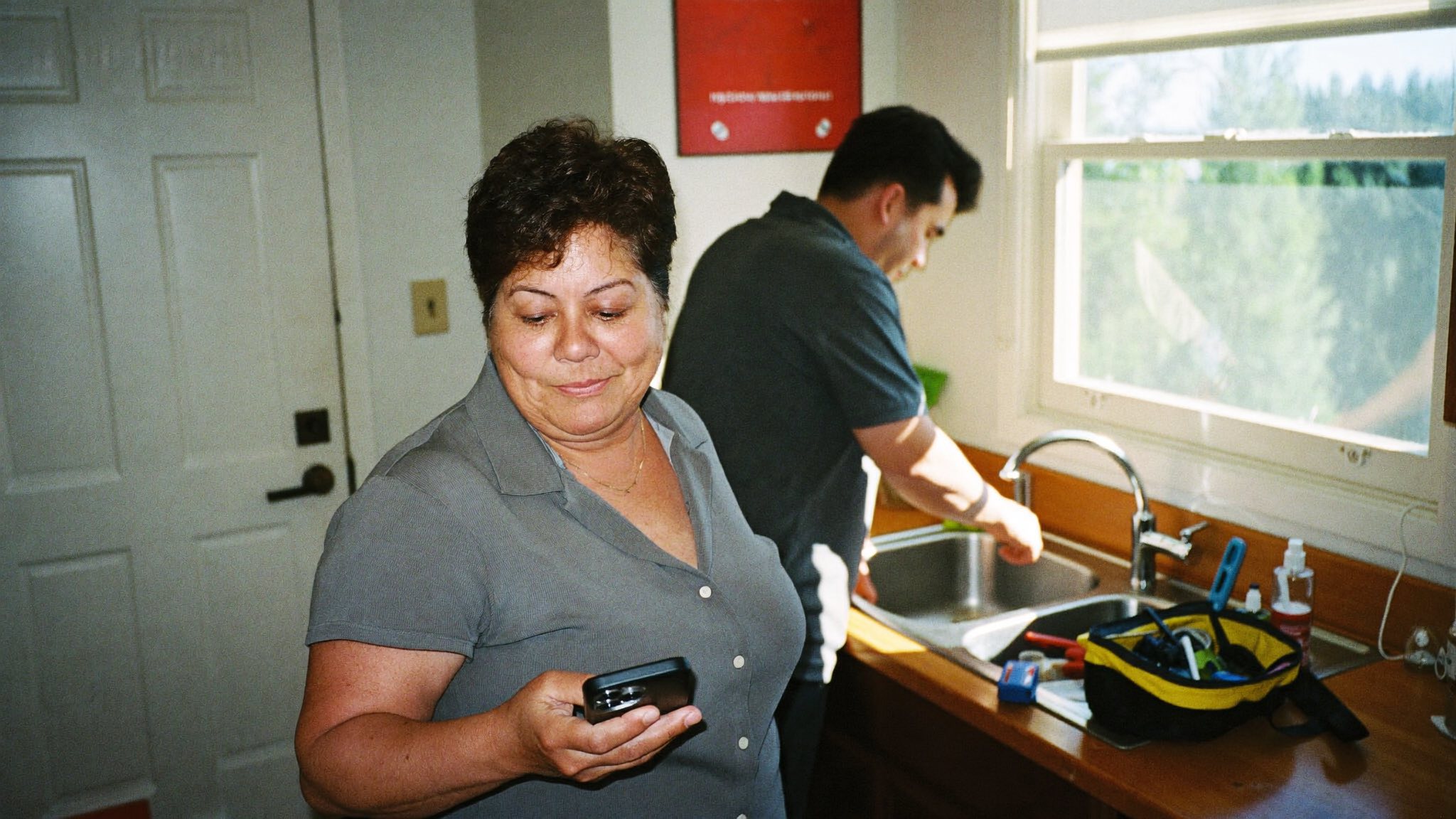 A homeowner smiling at her phone while a plumber fixes the kitchen faucet in daylight